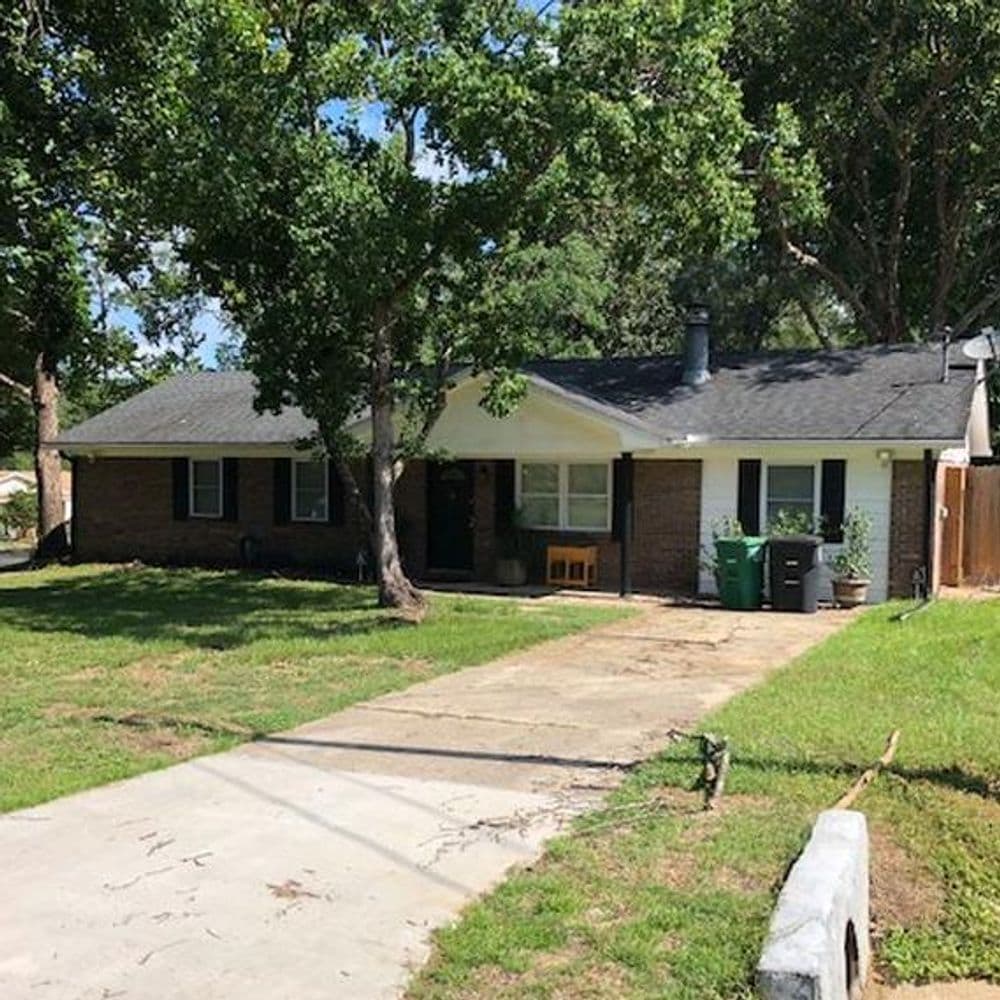 Single-story brick house with a green lawn, long driveway, and trees surrounding the property.