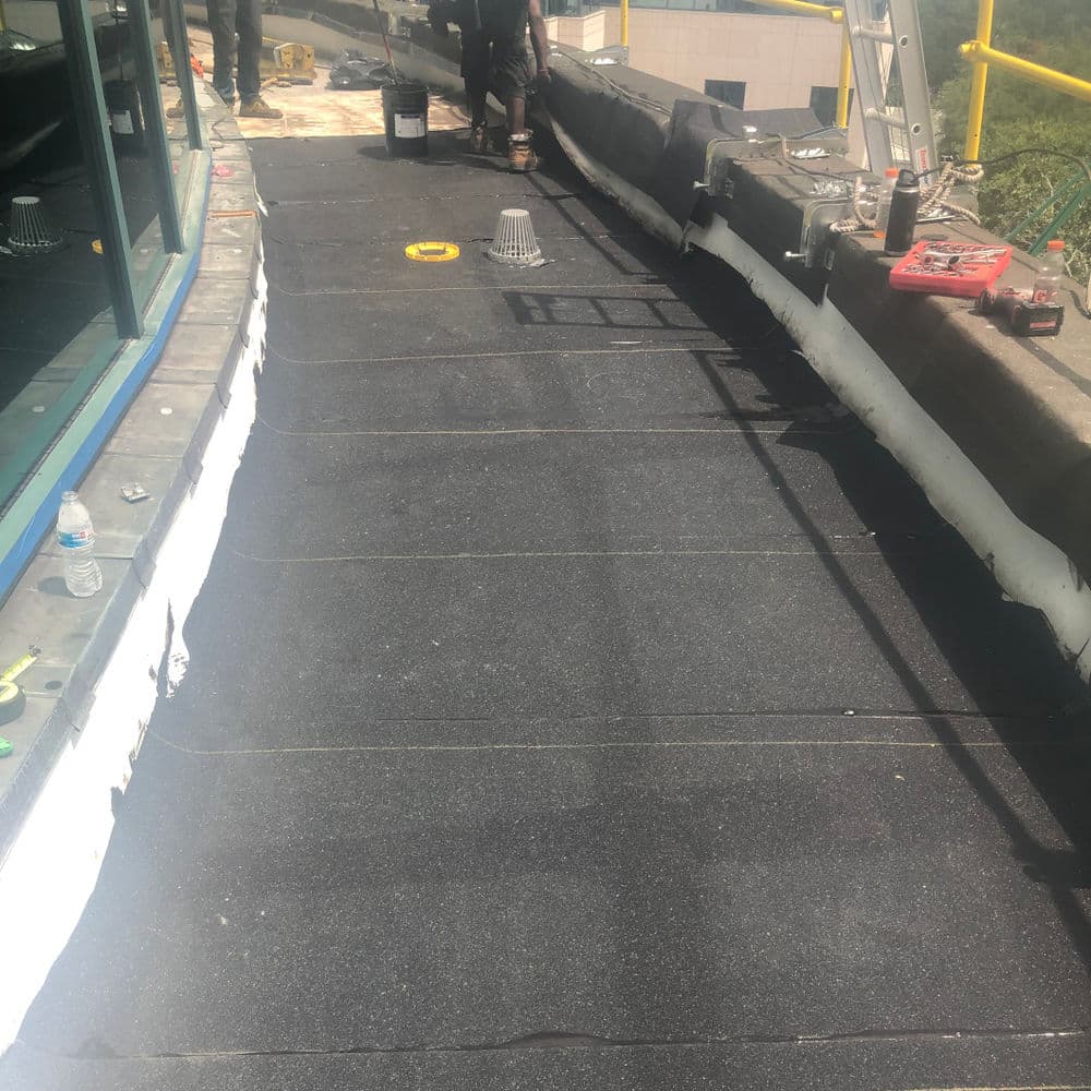 Workers installing black roofing material on a flat roof with tools nearby.