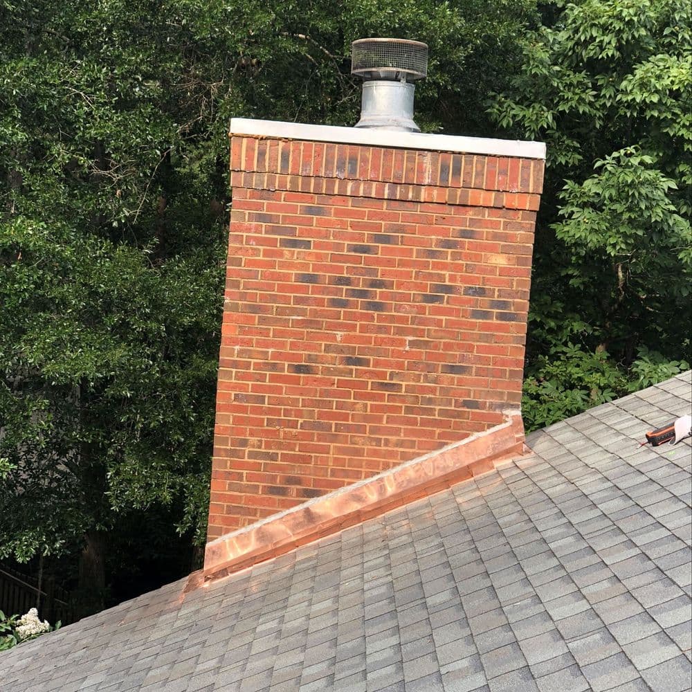 Brick chimney with metal cap on a sloped roof surrounded by trees.